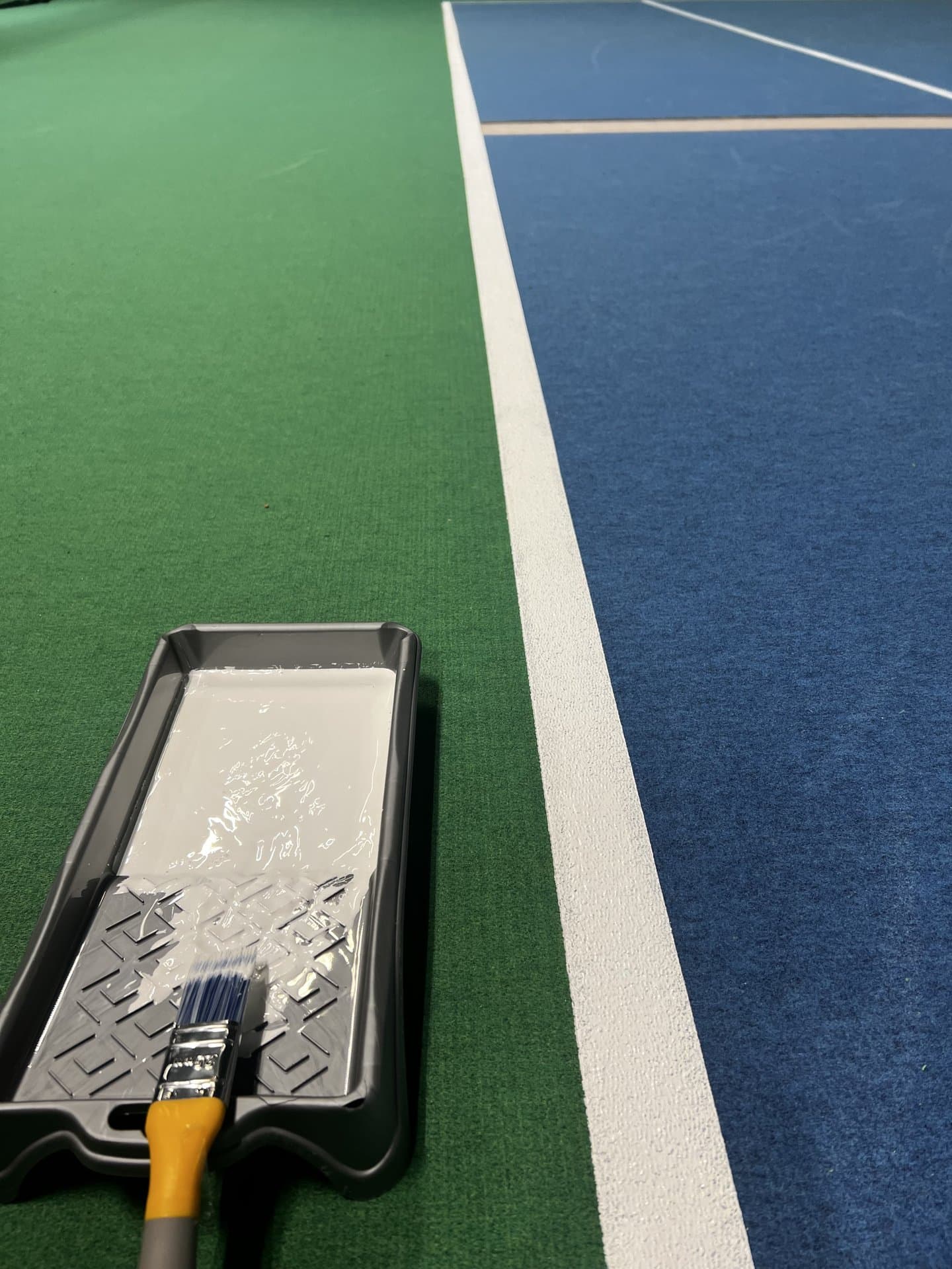 Närbild på målarbacke och pensel under linjering av sportgolv, gröna och blå ytor möts vid vit linje